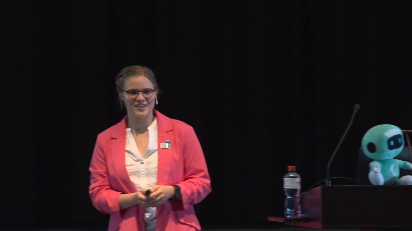 Carolin Neumann on stage at German UPA 2023, with the Bitsy plush robot sitting on the lectern